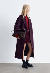 Langer lila Wollmantel, roter Strickpullover, burgunderfarbener Rock, schwarze Slipper und eine dunkelbraune Handtasche. Glatte Textur, maßgeschneiderter Schnitt.