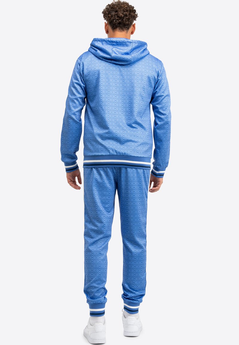 Blaues Hoodie und passende Hose aus strukturiertem Material mit geometrischem Muster, gerippten Bündchen und Kapuze. Trägt weiße Sneakers.
