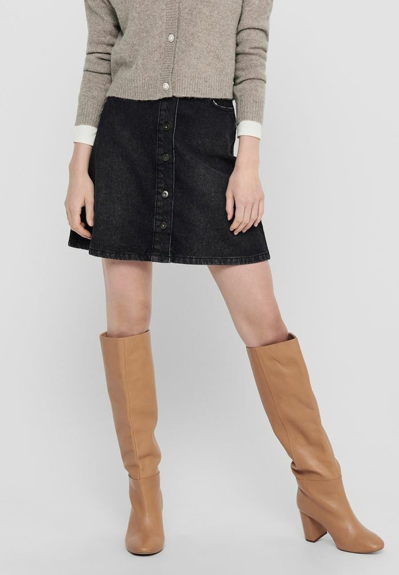 Femme portant un cardigan beige boutonné, une jupe en jean noire à boutons sur le devant, et des bottes hautes marron clair à talons, debout les jambes écartées.