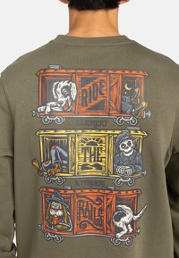Sudadera verde oliva con un diseño gráfico de animales de dibujos animados en vagones de tren, con llamativos acentos en naranja, amarillo y negro.