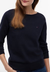 Donna che indossa un maglione blu navy a maniche lunghe con un piccolo logo rosso, bianco e blu sul petto sinistro e jeans blu.