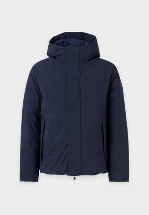 Homme portant une veste à capuche bleu marine, un t-shirt blanc col en V, un pantalon large marron et des baskets beige, debout devant un fond gris uni.