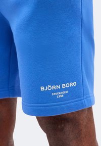 Μπλε σορτς που φοριέται από ένα άτομο, με λευκό κείμενο «BJÖRN BORG STOCKHOLM 1956» κοντά στο τελείωμα στο αριστερό πόδι.