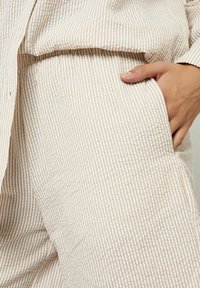 Conjunto de tela a rayas beige y blanco compuesto por una camisa con botones y pantalones de corte relajado, que destaca una superficie texturizada y un bolsillo lateral.