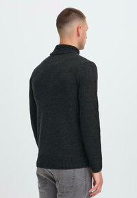 Man die met de rug naar ons toe staat, gekleed in een donkergrijze geribbelde coltrui en lichte grijze jeans tegen een effen lichte achtergron.