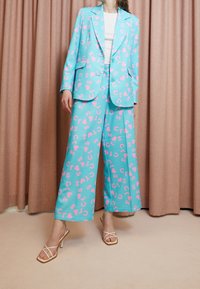 Zweiteiliges Anzugset aus türkisem Stoff mit rosa Herz- und Buchstabenmustern. Die Blazerjacke und die weit geschnittene Hose haben eine taillierte Passform und zwei Knöpfe.