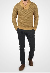 Strikket sweater i sennep med et tekstureret mønster, der har en sjal krave og knapdetaljer, parret med sorte bukser og brune sko.