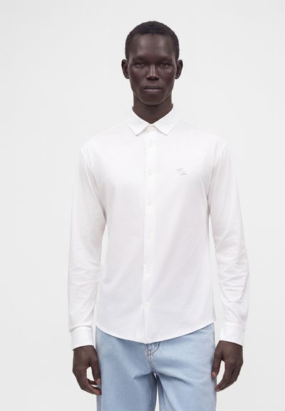Homme portant une chemise blanche unie à manches longues et un jean bleu clair, debout devant un fond blanc.