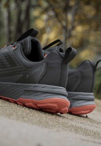 Paire de chaussures de trail Terrex noires et grises avec semelles orange placées sur un chemin de gravier avec des arbres d’automne flous en arrière-plan.