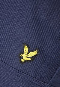 Mørkeblått stoff med en gul brodert fuglelogo, omkranset av svart. Teksturen er vevd med synlige sømdetaljer.