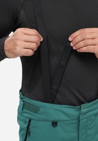 Grüne verstellbare Hose mit strukturierten schwarzen Hosenträgern. Der Bund verfügt über einen Klettverschluss und einen Reißverschluss vorne.