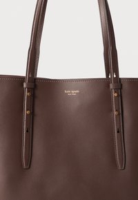 Brown leather tote bag with dual straps, featuring gold logo and hardware accents. Smooth texture with visible stitching and cutouts.
