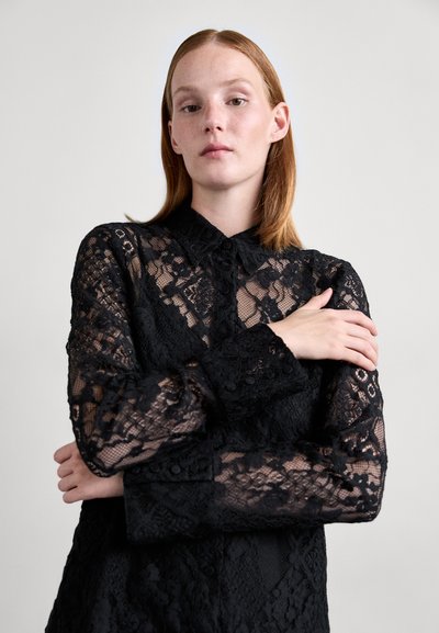 Blouse en dentelle noire avec un col à boutons, des manches longues et des motifs floraux élaborés. Présente un tissu transparent et des détails texturés sur l'ensemble.