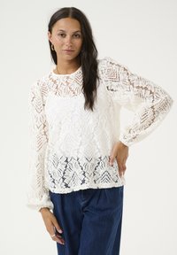 White lace blouse with a floral pattern, long puff sleeves, and a round neckline, paired with dark denim pants. Intricate textured design.