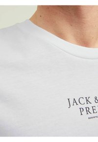 Bílá bavlněná tričko s kulatým výstřihem. Obsahuje potištěné logo "JACK & PRE" tmavým písmem na oblasti hrudi.