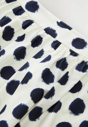 White fabric skirt featuring large navy blue polka dots. The texture is smooth with gathered pleats at the waistband.
