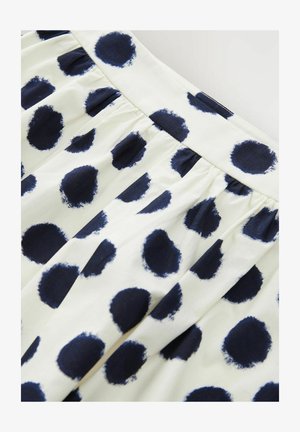 White fabric skirt featuring large navy blue polka dots. The texture is smooth with gathered pleats at the waistband.