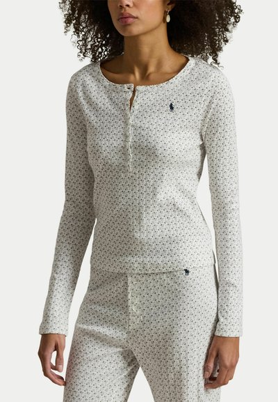 Vrouw draagt een wit langärmelig henley-shirt en bijpassende broek met een klein zwart bloemenpatroon en geborduurde marineblauwe polo-spelerlogo's.