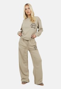 Blonde vrouw in een beige oversized sweatshirt en wijde broek met "Brooklyn New York" en luipaardprint, staande met handen in haar zakken.