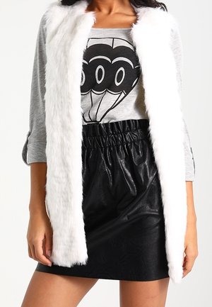 Woman wearing a white faux fur vest over a gray graphic top and a high-waisted black leather skirt, standing with one hand relaxed.