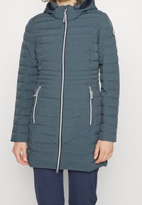 Polstret jakke i mørk teal med horisontal quiltning, to sidelommer med lynlås, en fuld frontlynlås og en hætte. Glat tekstur og letvægtsdesign.