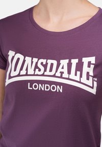 Purple cotton t-shirt featuring a large white logo that reads "LONSDALE" with "LONDON" below in smaller text. Round neckline, smooth texture.