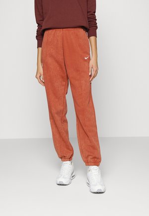 Person wearing rust-colored Nike sweatpants and white sneakers, standing against a plain light background.