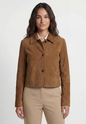 Frau trägt eine braune Wildlederjacke mit Knöpfen über einem beigen Hemd mit Kragen und beigen Hosen, steht vor einem schlichten weißen Hintergrund.