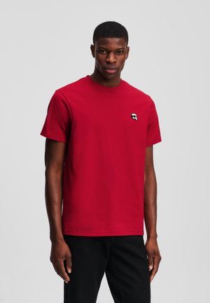 T-shirt en coton rouge avec un col rond, des manches courtes et un petit logo noir et blanc sur la poitrine. Texture lisse, coupe décontractée.
