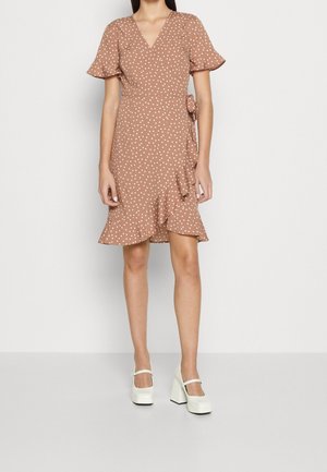 Robe portefeuille à pois marron avec des manches courtes à volants, un lien sur le côté et un ourlet à volants ; associée à des chaussures blanches à talons épais.