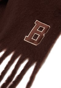 Bufanda de lana marrón con extremos con flecos y una letra "B" bordada en marrón y crema en un lado, resaltando su suave textura.