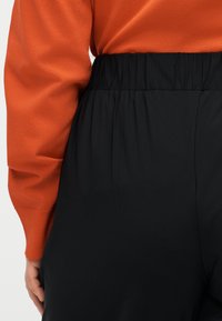 Άτομο που φοράει μαύρο παντελόνι ψηλόμεσο με ελαστική μέση και πορτοκαλί μπλούζα μακρυμάνικη, με το χέρι ακουμπισμένο στο πλάι.