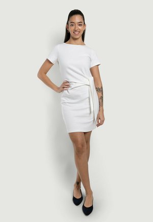 Donna con coda di cavallo liscia, vestito bianco al ginocchio con cintura, tacchi neri e tatuaggio floreale sul braccio, in piedi con sicurezza su sfondo semplice.