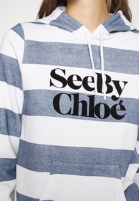 Personne portant un sweat à capuche bleu et blanc à rayures avec le logo "See By Chloé" imprimé en noir sur la poitrine.