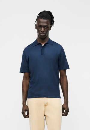 Fiatal férfi sötét, fonott hajjal, sötétkék rövid ujjú galléros pólóban és világos bézs nadrágban, egyszerű háttér előtt állva.