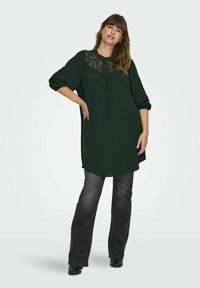 Tumši zaļš tunika ar mežģīņu apdari ap kaklu, brīvs piegriezums un trīs ceturtdaļu piedurknes, apvienota ar tumšām platām džinsām un melnām zābakiem.