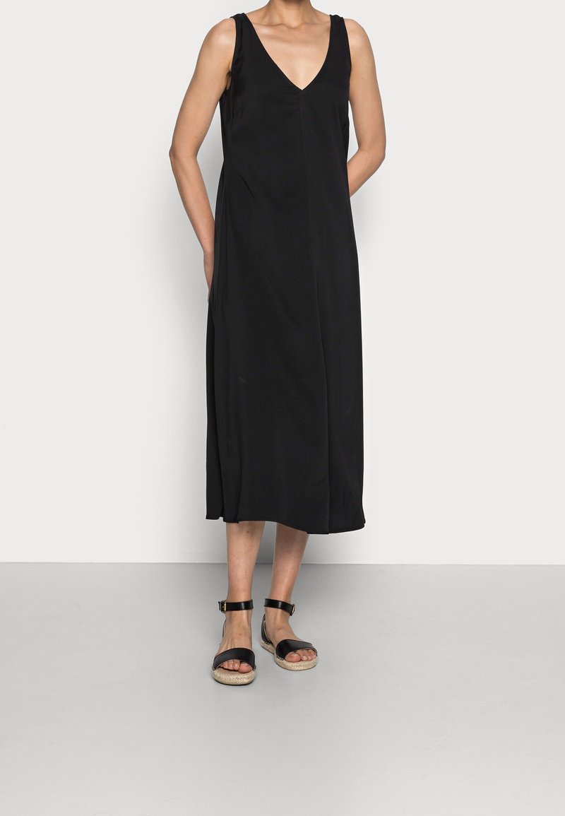 Femme portant une robe midi noire sans manches avec un décolleté en V profond et des sandales noires à bride cheville, debout devant un fond clair.
