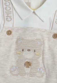 Salopette beige en coton avec motif d'ours brodé. Comprend un bouton en bois, des accents bleu clair, et un col blanc assorti. Texture douce.
