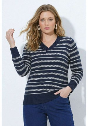 Kvinde med bølget blondt hår iført store hoop-øreringe, en marineblå og hvid stribet V-hals sweater og blå bukser, poserende med den ene hånd hævet.