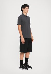 Ungdommelig mann stående i en mørkegrå polo skjorte, svarte knelende shorts, svarte sokker og svarte joggesko mot en ensfarget hvit bakgrunn.