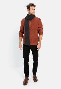 Verbrennter orangefarbener Strickpullover mit einem Viertelreißverschluss, kombiniert mit einem dunkelgrauen gerippten Schal, schwarzen Jeans und braunen Lederschuhen.