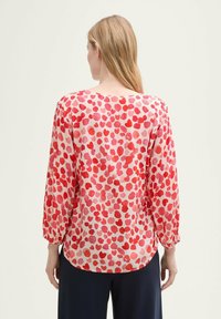 Blusa leggera a maniche lunghe in tessuto rosa con motivo a cuori in diverse sfumature di rosso. Presenta un collo tondo e polsini elasticizzati.