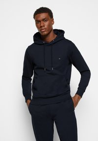 Felpa con cappuccio blu navy realizzata in tessuto morbido, con tasca a marsupio frontale, corde regolabili e un piccolo logo sul petto.