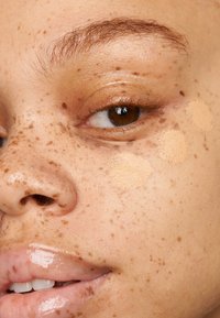 Nahaufnahme eines Gesichts mit einer sommersprossigen Haut. Cremige Foundation-Proben in verschiedenen Hauttönen auf der Wange, glänzende Lippen und braune Augen.