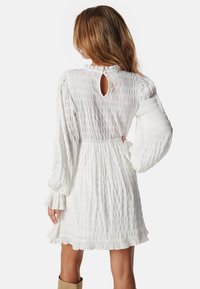 Biała sukienka z teksturowanej tkaniny, z długimi bufkami i otworem w kształcie klucza na plecach. Rękawki są marszczone dla dodatkowego akcentu.