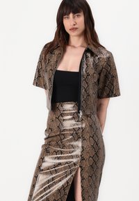 Tenue à motif peau de serpent composée d'un haut zippé ajusté et d'une jupe crayon avec une fente sur le côté, le tout en tons de brun métallique et avec une texture brillante.