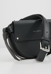 Zwarte leren crossbodytas met een klepsluiting, zilverkleurige metalen details en een verstelbare riem. Heeft een gladde textuur en een minimalistisch ontwerp.