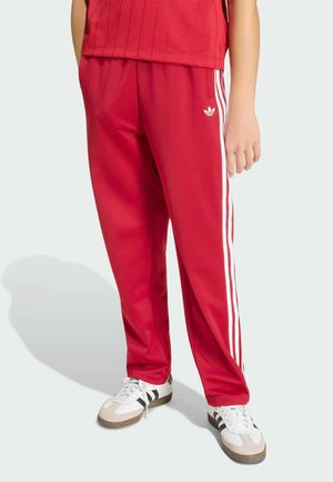 Persoon die rode Adidas trainingsbroek met witte strepen en witte sneakers draagt, staand op een lichte achtergrond.