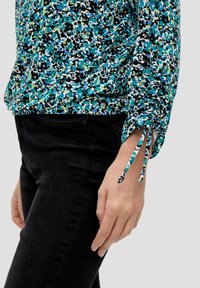 Blumenmuster, mehrfarbige Bluse mit einem blau, grün, schwarz und weißen Muster. Verfügt über geraffte Ärmel mit Kordelzügen. Kombiniert mit schwarzen Hosen.
