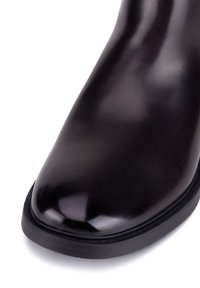 Černé kožené kotníkové boty s lesklým povrchem, zaoblenou špičkou a nízkou gumovou podrážkou. Mají bezšvový design s jemnými švy.